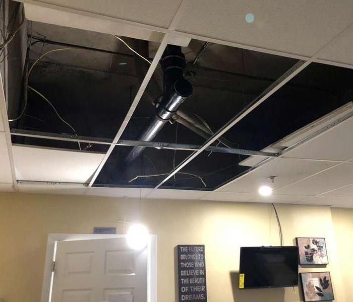 Open tiles in ceiling show broken water pipe in room with tv and pix on wall